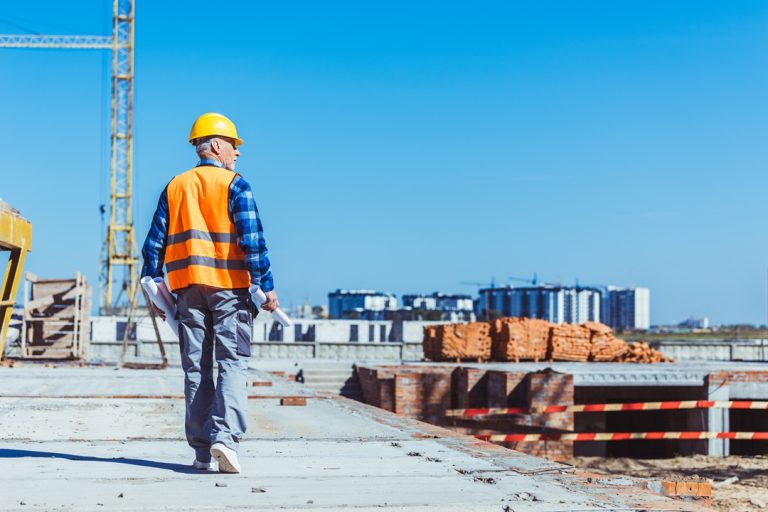 Builder in reflective vest and hardhat walking across a construction site with rolls of plans in his Builder in reflective vest and hardhat walking across a construction site with rolls of plans in his